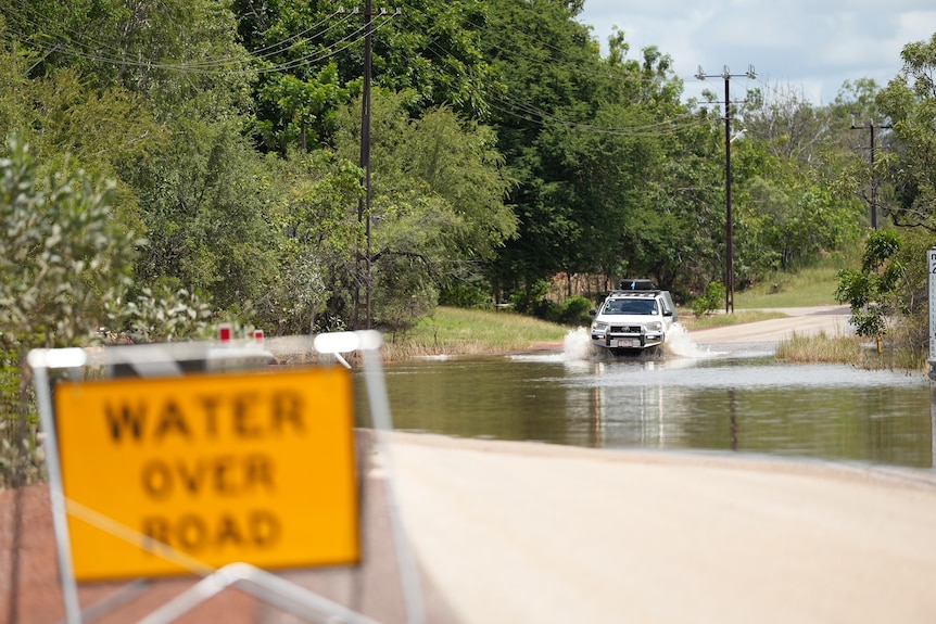 A white SUV in focus in background driving through flood waters, with 'water over road' sign in foreground