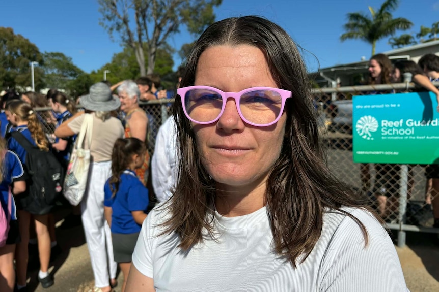 A dark-haired woman in glasses stands near a crowd of people outside a high school.