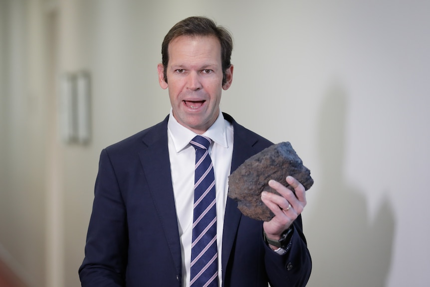 A man in a dark suit holds a lump of coal while standing in a corridor.
