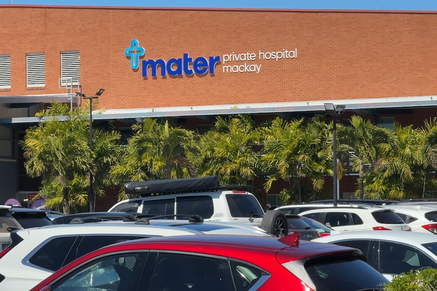 A cross symbol and the name "Mater Private Hospital Mackay" lettered on the side of a brick building behind a car park.