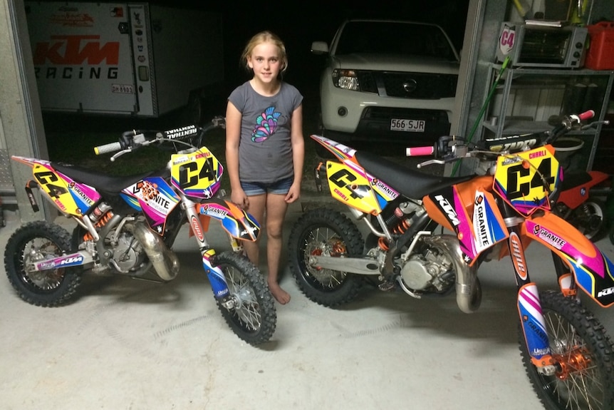 A young girl standing in between two dirbikes.