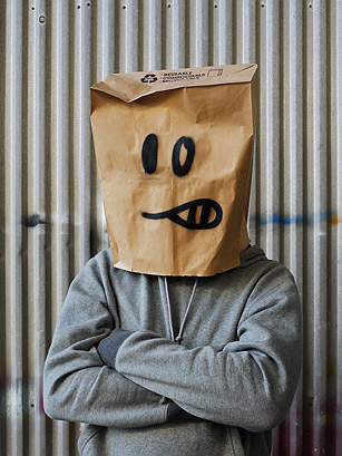 A photo of a man in a grey hoodie wearing a paper bag over his head with a cartoonish grimace drawn on with a marker.