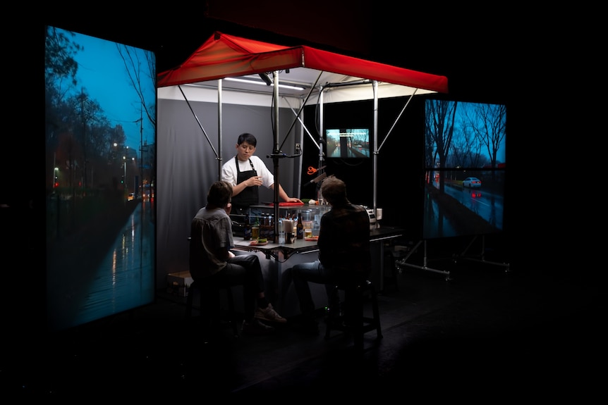 Man on stage inside food stall talks to two people