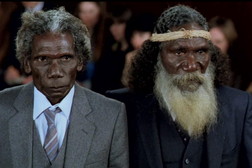 Two Aboriginal men stand looking serious, both wear suits.