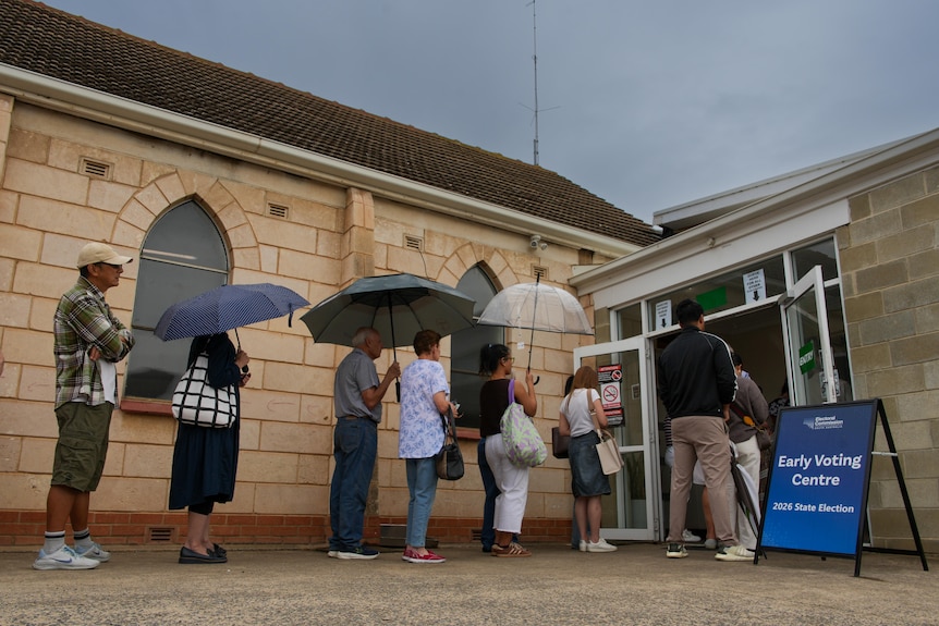 A queue of people some using umbrellas outside a stone building with a sign saying Early Voting Centre