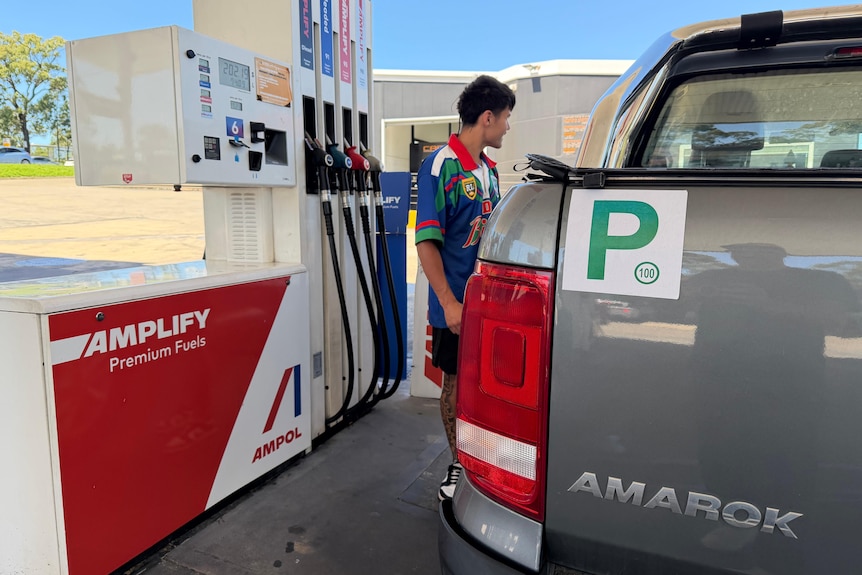 A young, dark-haired man fills up adual-cab ute at a petrol station on a sunny day.