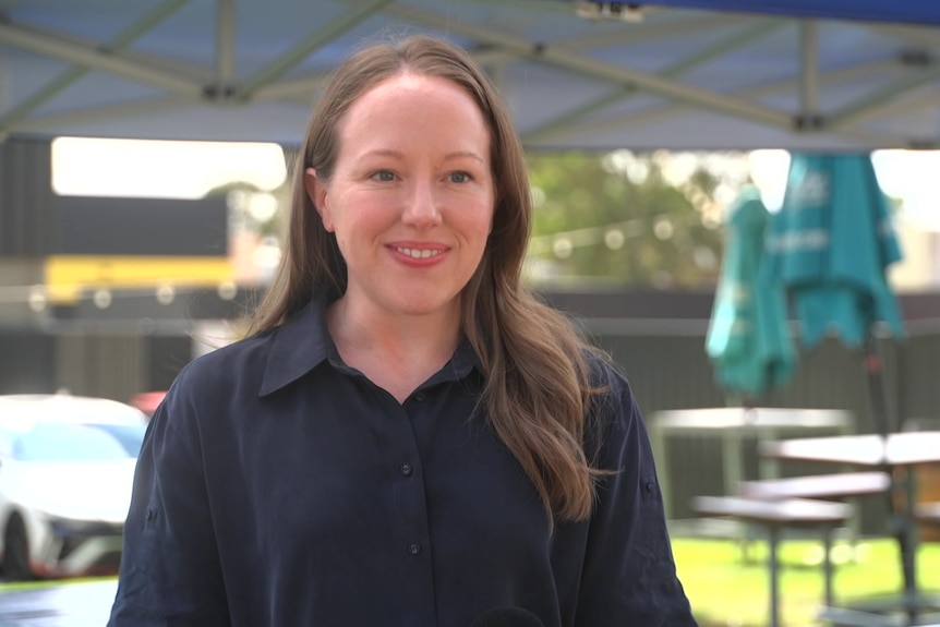 A smiling woman with long brown hair wearing a black shirt stands outside with tables and chairs behind her