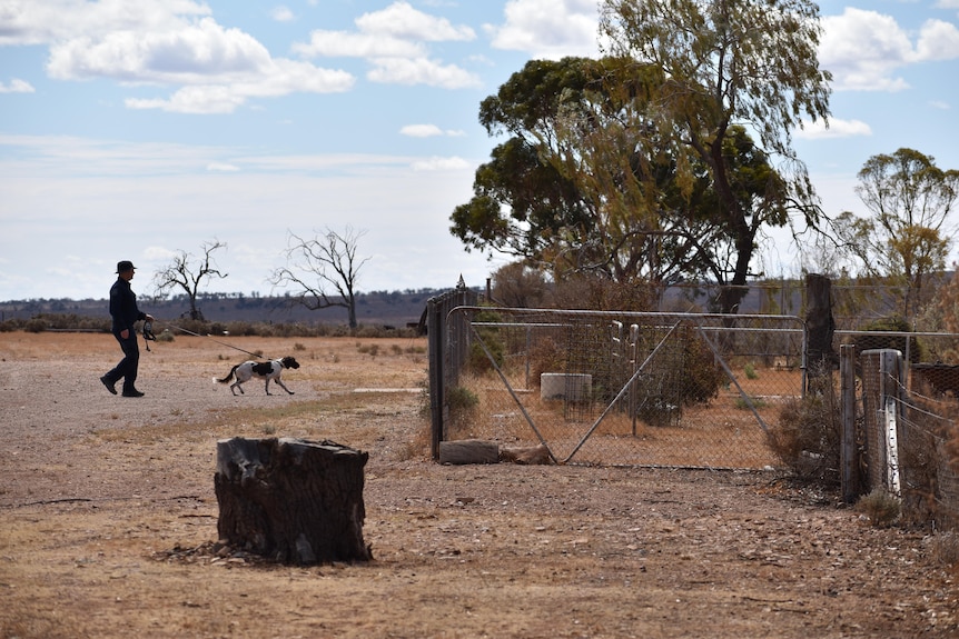 A police officer with a dog on a leash walking through the gates of a rural property