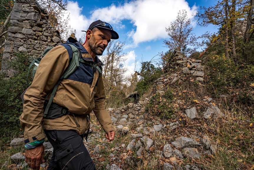 A rugged-looking man dressed for hiking, walking a trail.