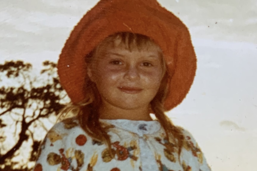 A young blonde girl smiles at the camera wearing swimwear and a floppy red sunhat.