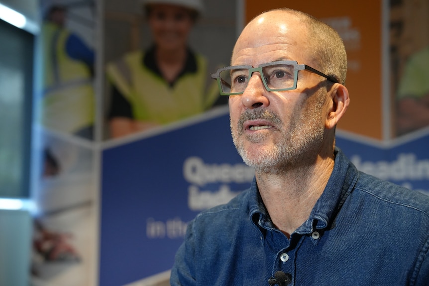 a man with square glasses and short grey hair speaks in front of a building sign