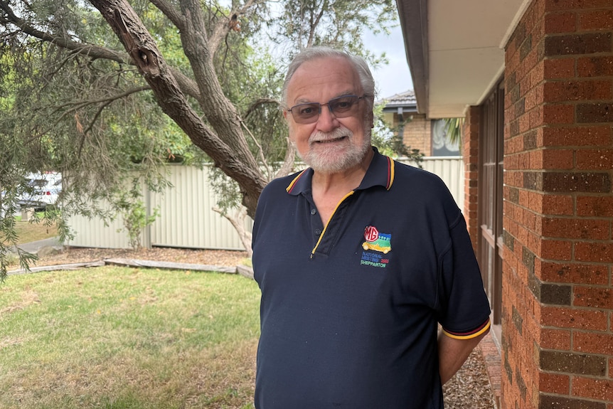 A man with white hair, beard and glasses in a blue polo shirt stands in front of a brick house with his hands behind his back.