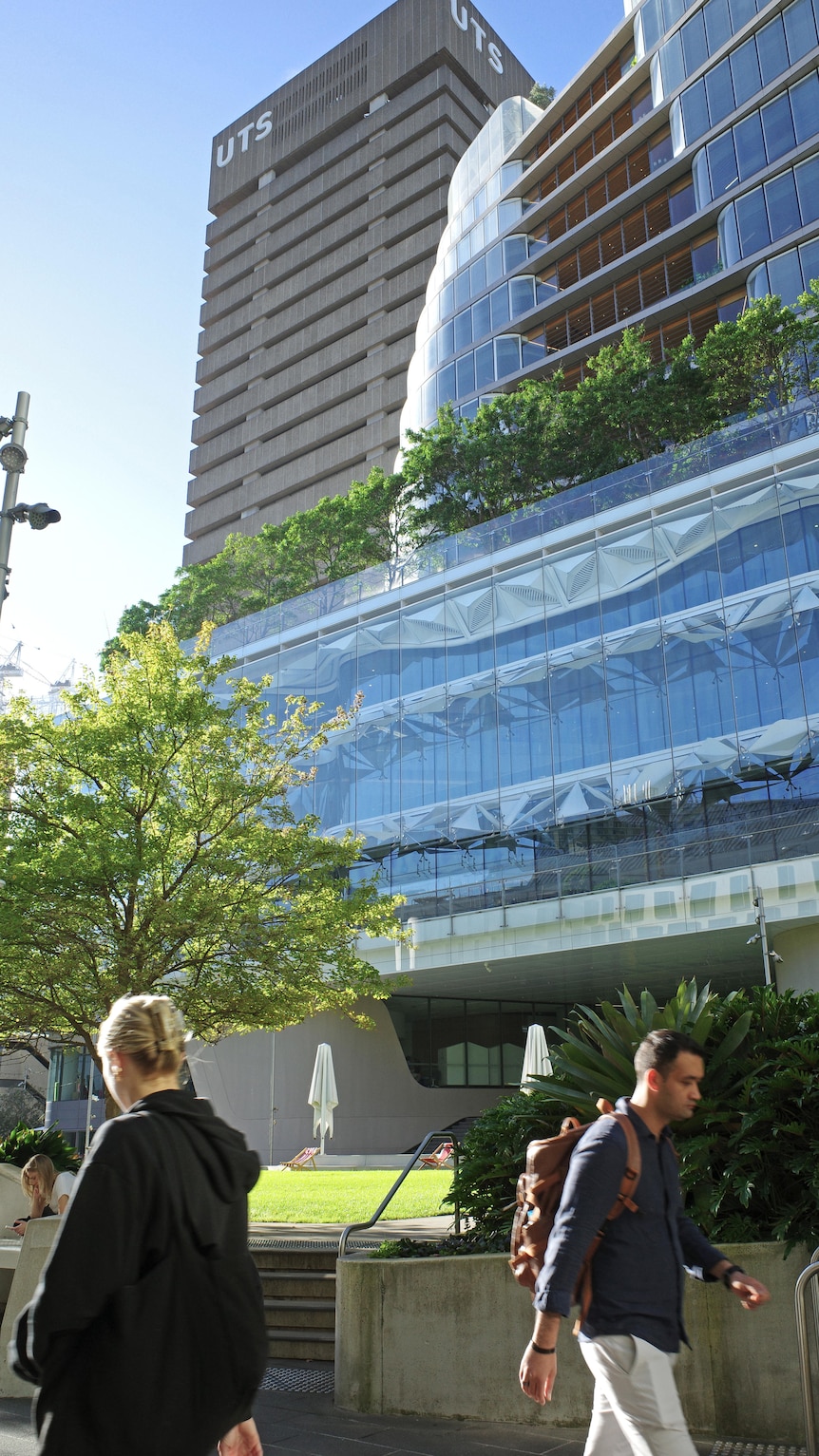 Two people pass is each other, behind them loom two large buildings, one a brutalist tower, the other is modern glass.