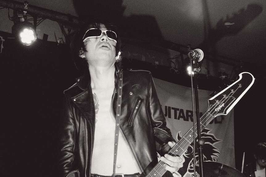 Black and white photo of a Japanese guitarist wearing a leather jacket, no shirt and sunglasses inside on a stage