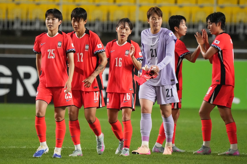A group of Korean soccer players in red, as well as one goalkeeper in grey, walk on a soccer field.