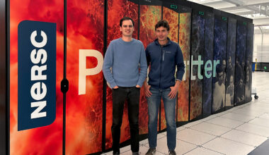 Two researchers stand in front of a colorful supercomputer. A NERSC logo and some of the letters of