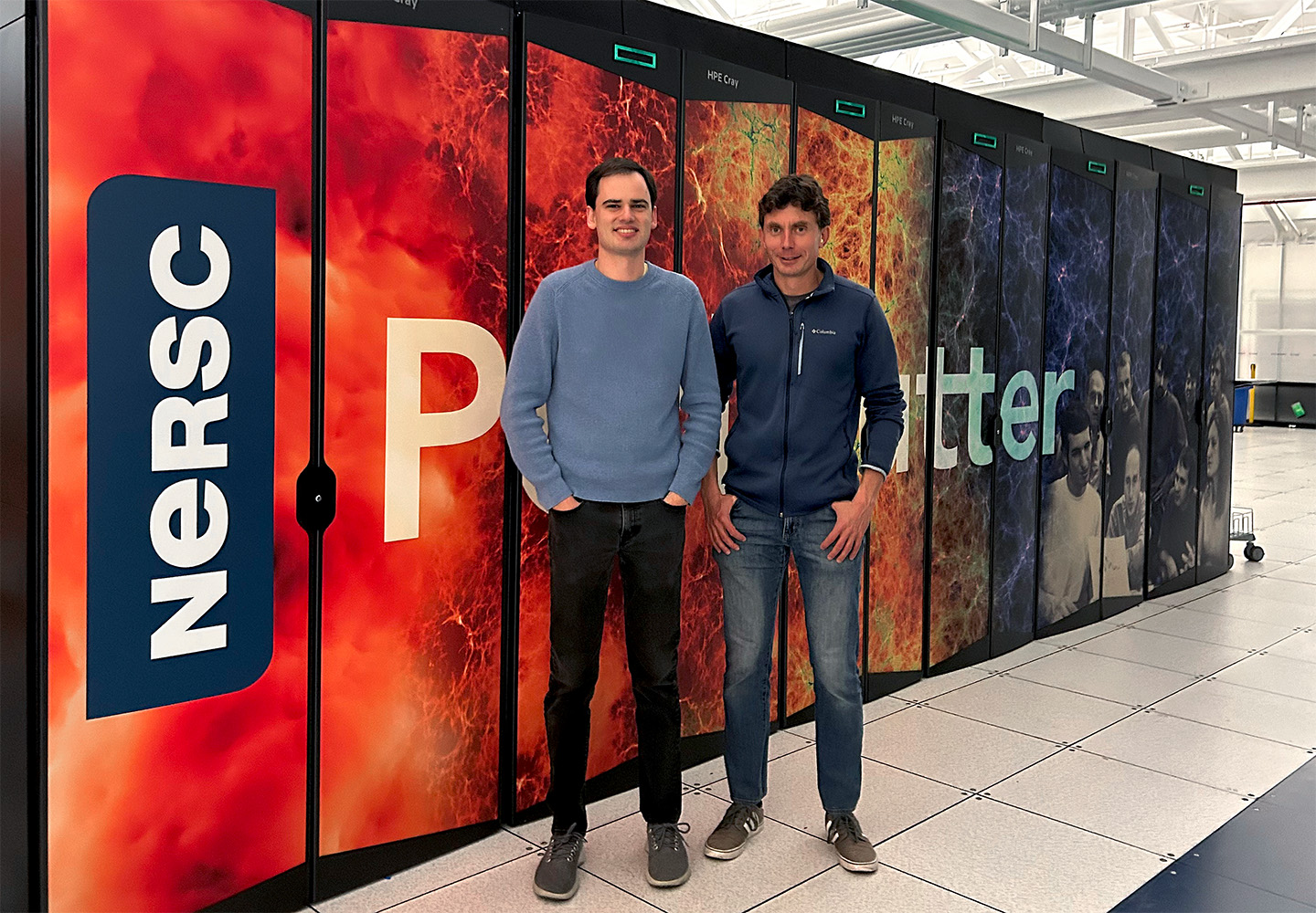 Two researchers stand in front of a colorful supercomputer. A NERSC logo and some of the letters of