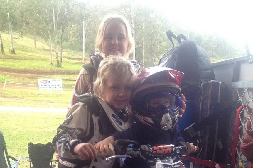 Three young kids dressed in motocross gear surround a dirtbike.