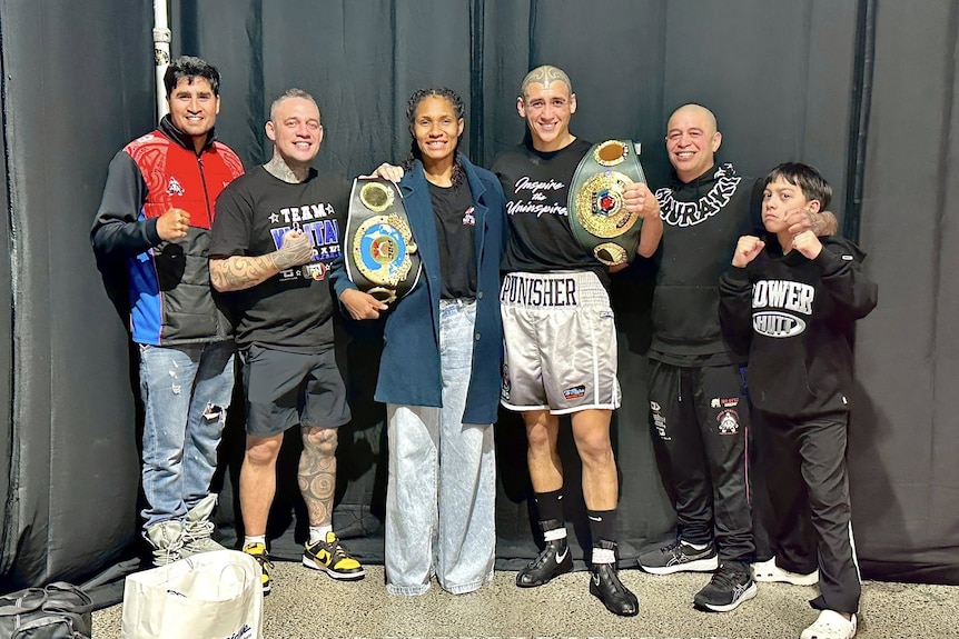Two champion fighters with their belts stand with a group of people backstage at a fight event and pose for a photo 
