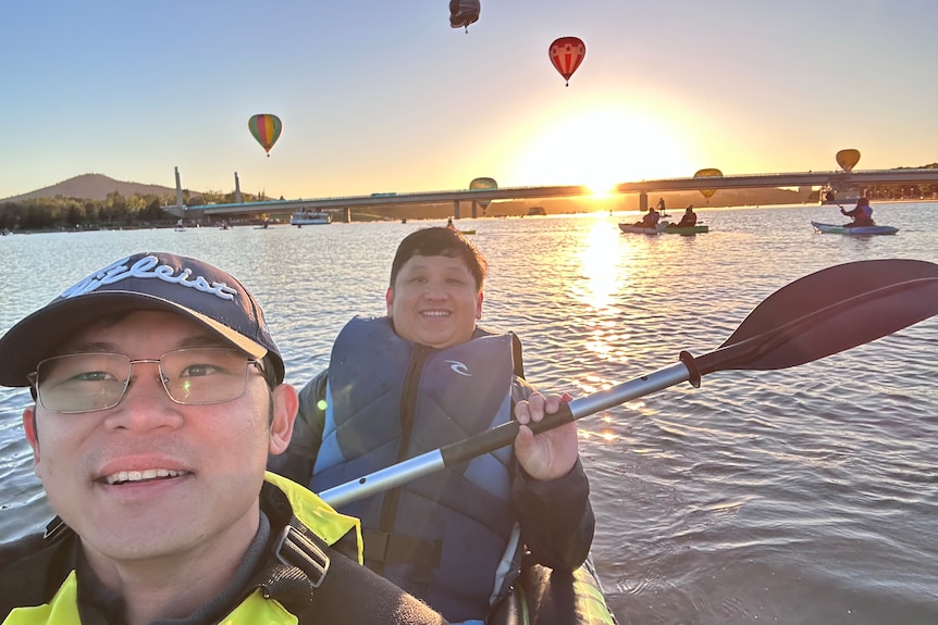 Two men on a kayak with hot air baloons in the background. 
