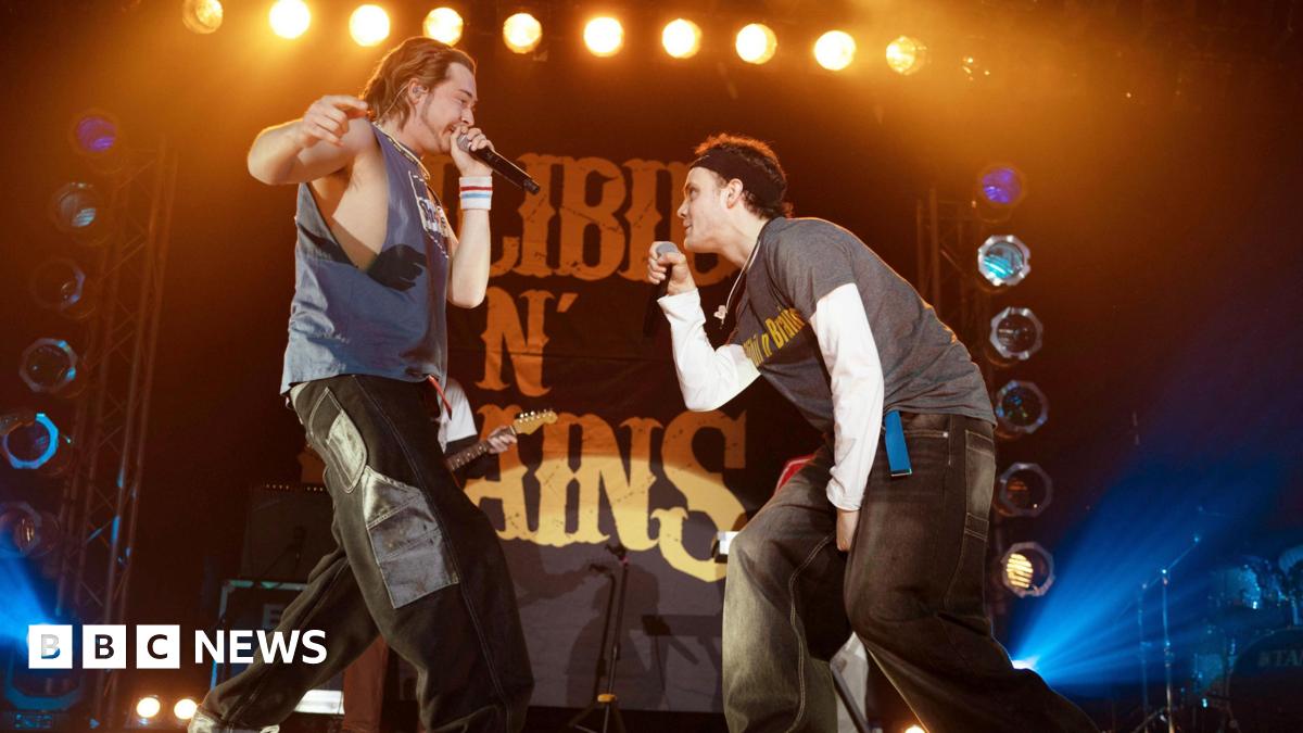 Two rappers in late 90s/early noughties clothing performing on stage with a backdrop of bright lights and a large sign that reads "Silibil N' Brains"
