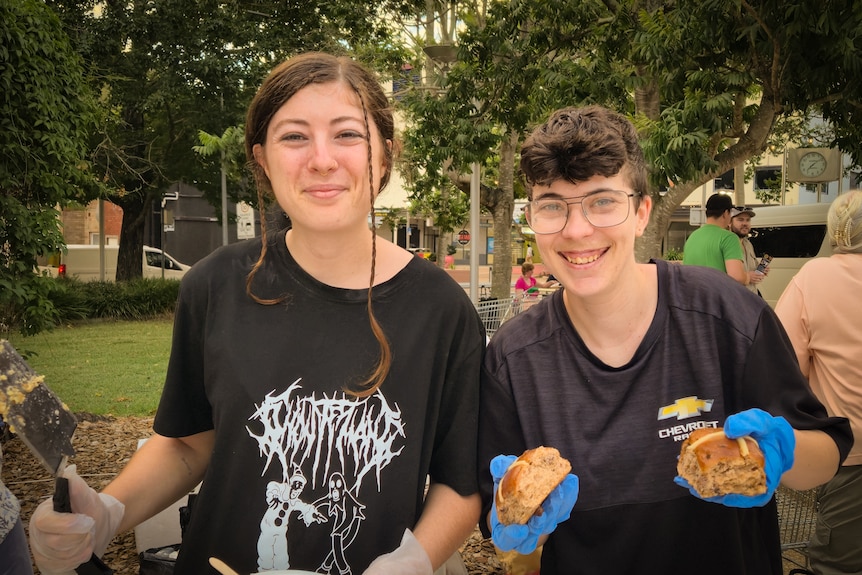 Two people wearing black holding food and serviing utensils behind a table of food trays.