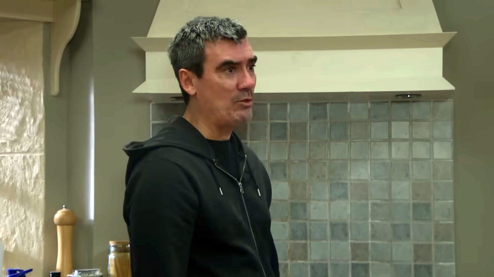 emmerdale, cain dingle in his tiled kitchen wearing a black hoodie and looking angry