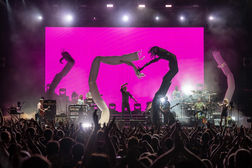 A cheering crowd watches Jarvis Cocker, 62, an arm in the air, and band Pulp on stage, flanked by air dancers.