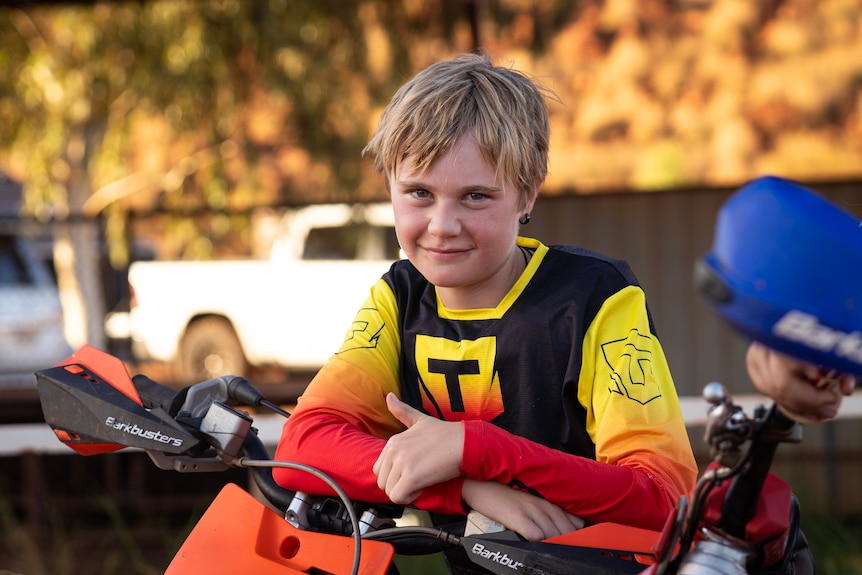 A young boy with blond hair leans up against the handlebars of his motocross bike.