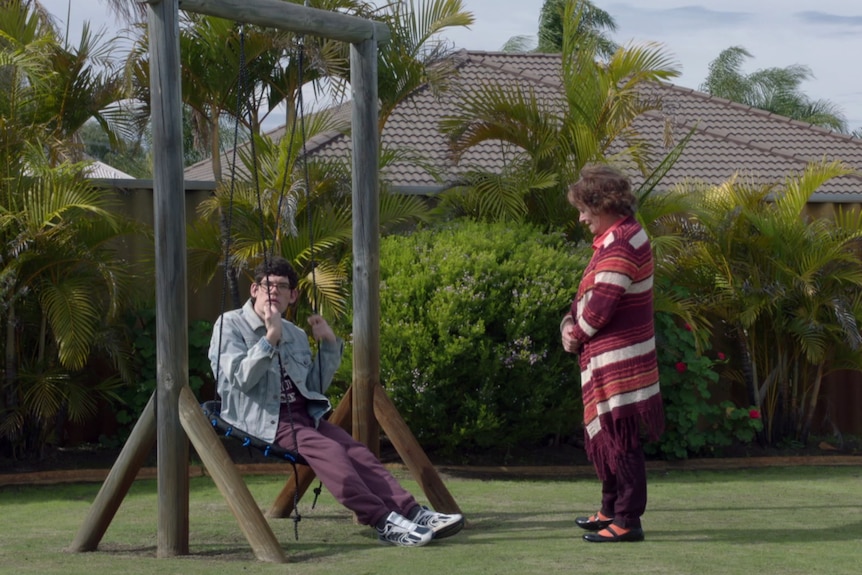 A man in jean jacket and long pants sits on an outdoor swing. His mother stands next to him dressed in tones of red 