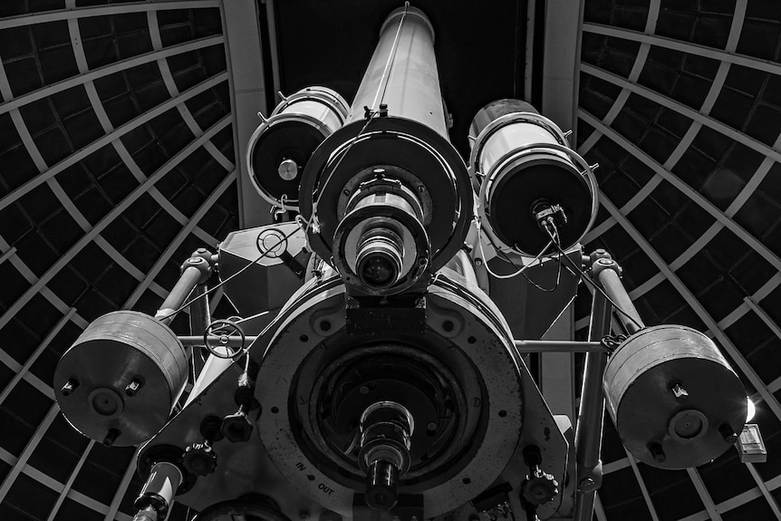 A multi-lens telescope pointing up to space, photographed from behind, in an observatory room.