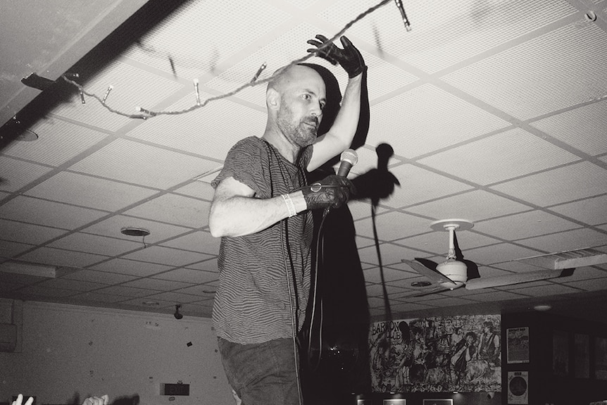 Black and white photo of man with microphone standing atop lively audience in an indoor music venue