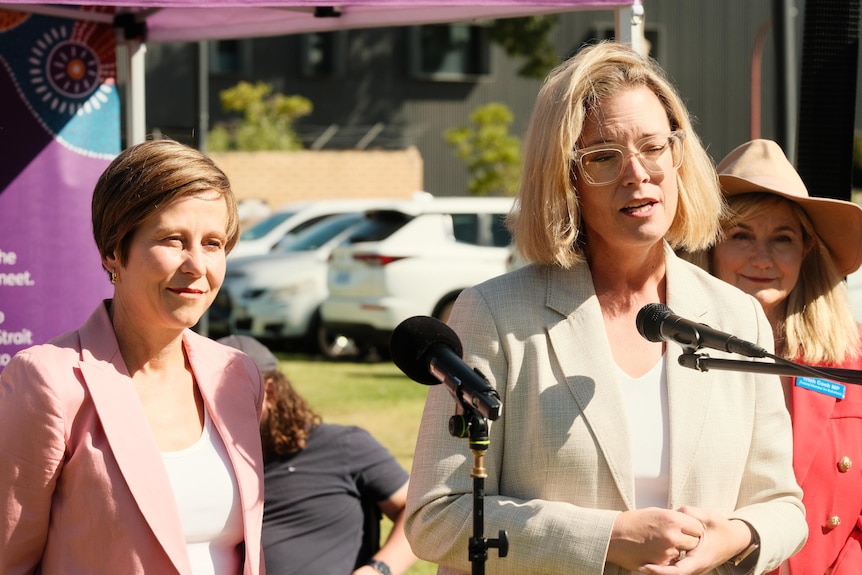 Image of Hannah Beazley speaking into a microphone. She wears glasses and a suit jacket.