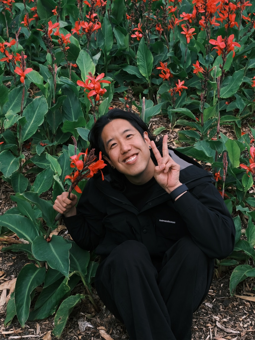 Harry Jun, a young Korean Australian, crouches beside a bush of red flowers, smiling and giving the peace sign.