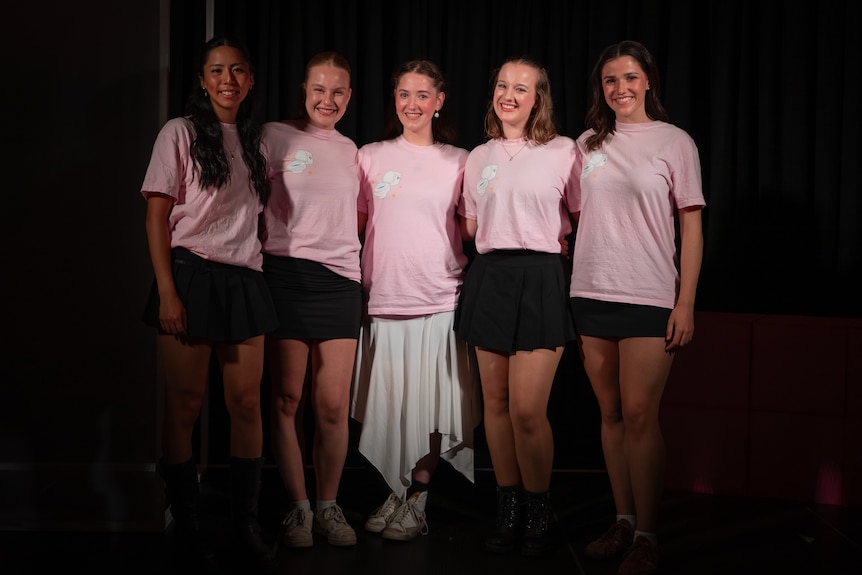Five young women wearing matching pale pink t-shirts stand in a line, smiling, against a darkened backdrop.