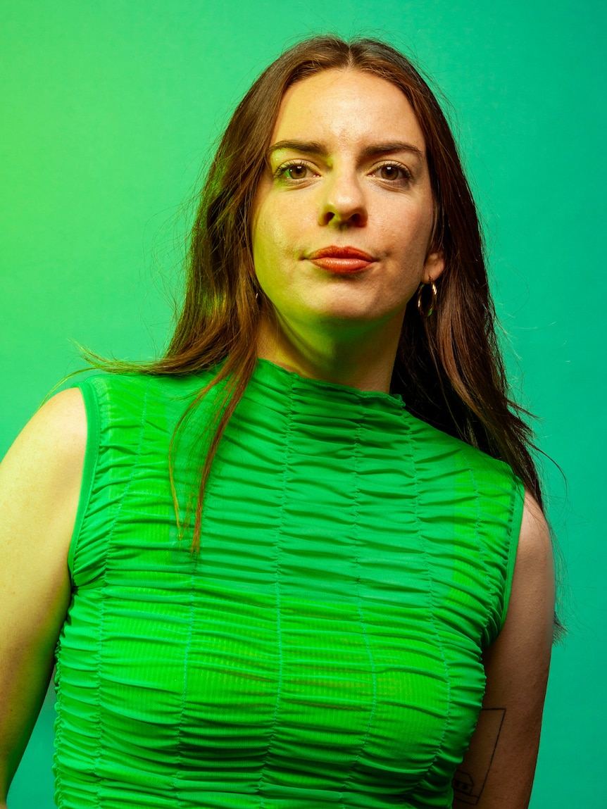 A portrait of Emma Holland, early 30s, with long brown hair, in a green sleeveless blouse, with a defiant expression.