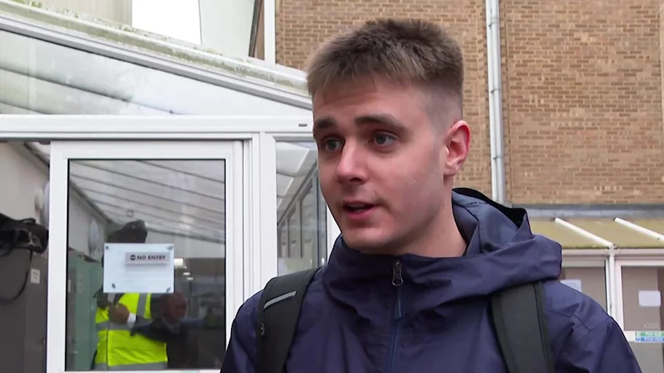 A head and shoulders image of James Thompson. He is wearing a navy coat over which he has two black rucksack straps. He is looking directly past the camera and standing in front of a conservatory-like entrance to a university building.