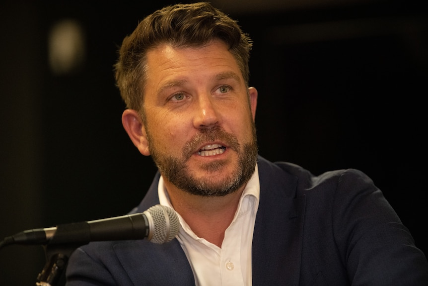 Close up photo of Moran speaking into a microphone, wearing white collared shirt with dark suit jacket