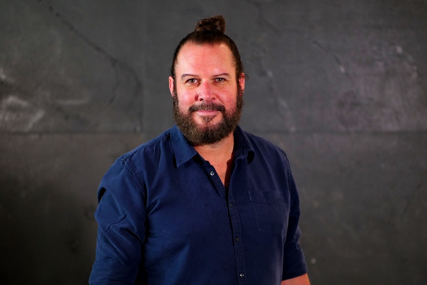 Man with brown hair in a bun and brown beard smiling in a navy blue shirt.
