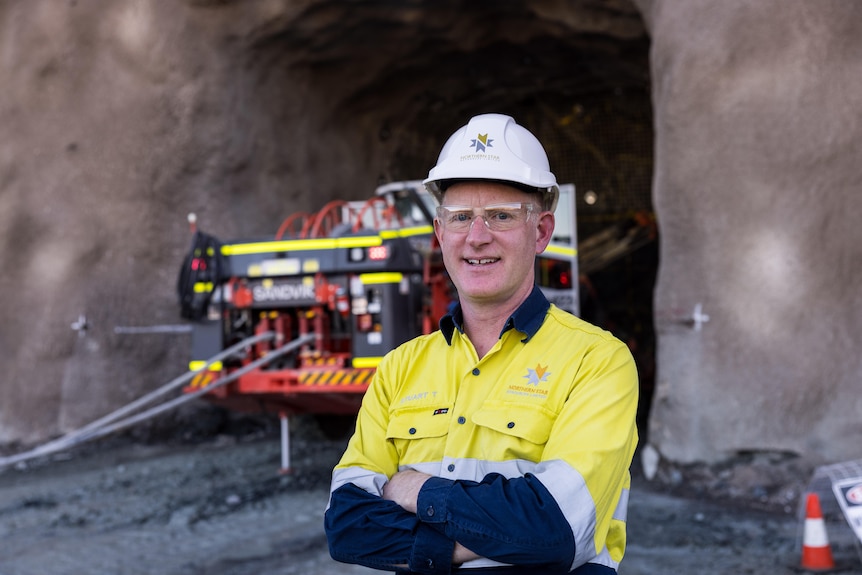 A man in high-vis and PPE stands in front of a machine at the mouth of a tunnel on a mine site.