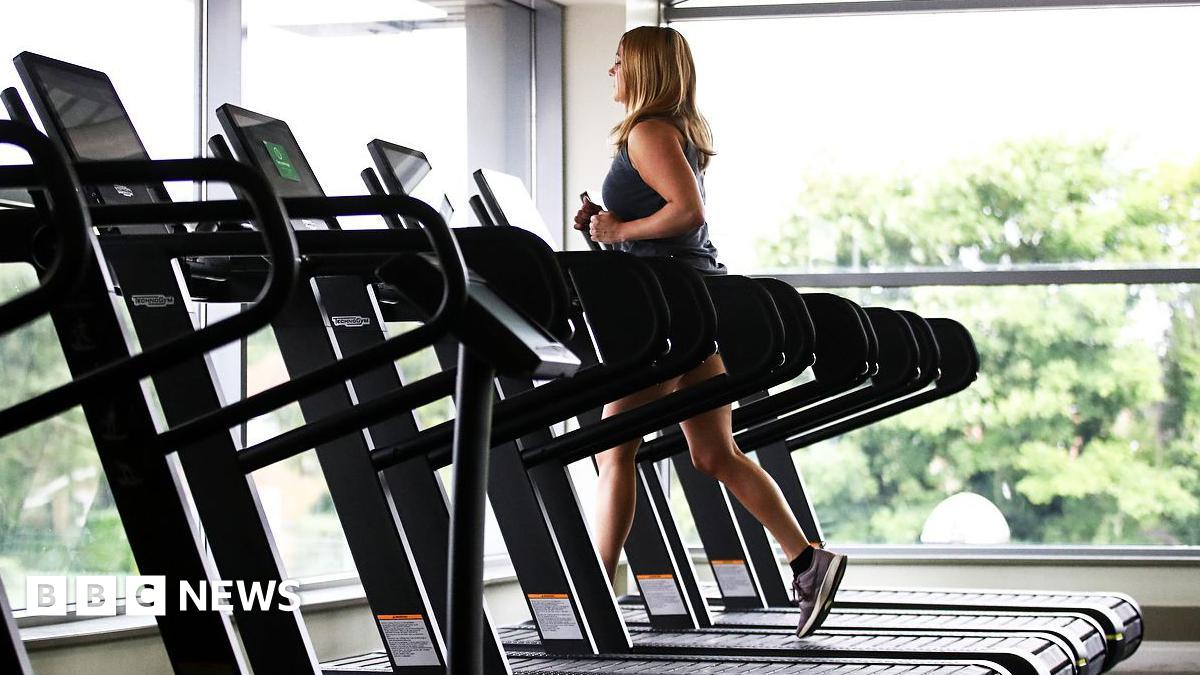 A woman wearing gym gear runs of a treadmill near large glass windows with trees outside. There are several empty treadmills next to her.