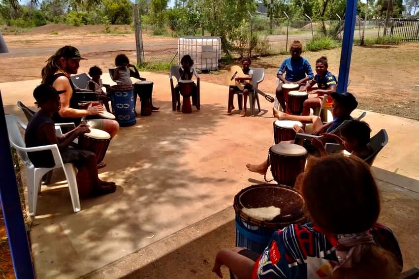 A man sitting in a circle with children, playing drums and native instruments. 