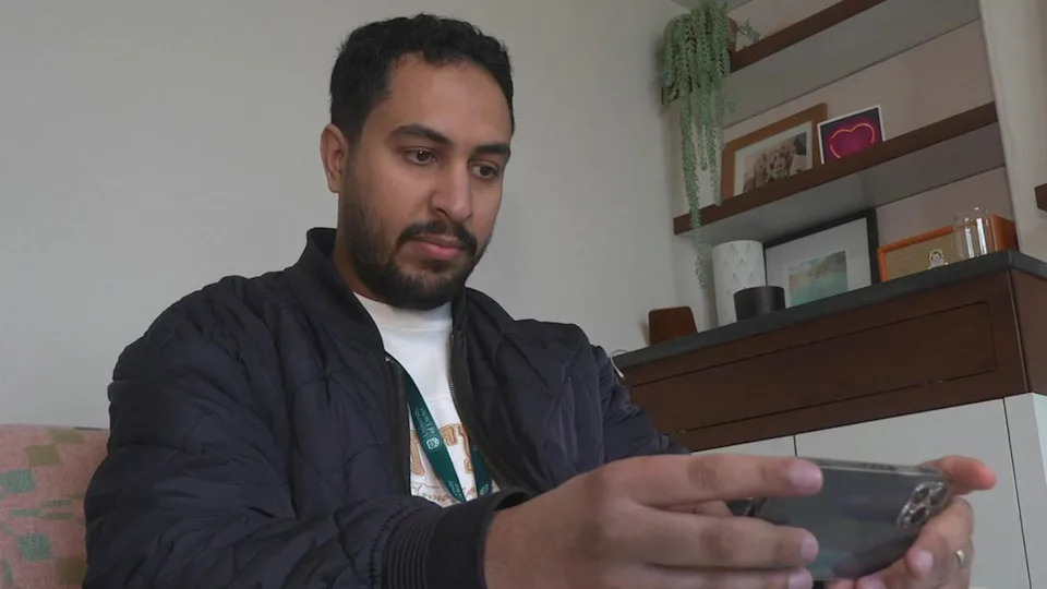 A man with short dark hair is sat on a sofa looking at virtual reality on his phone. We cannot see the phone screen, but he is monitoring the VR that the participants see on a headset. 