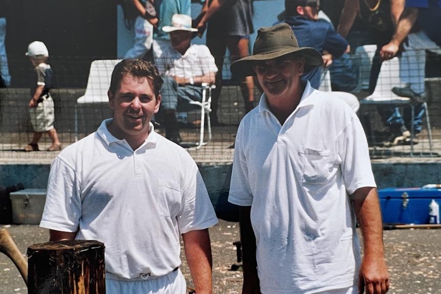 father and son in white tops and pants stand together, with the son holding an axe next to a log.