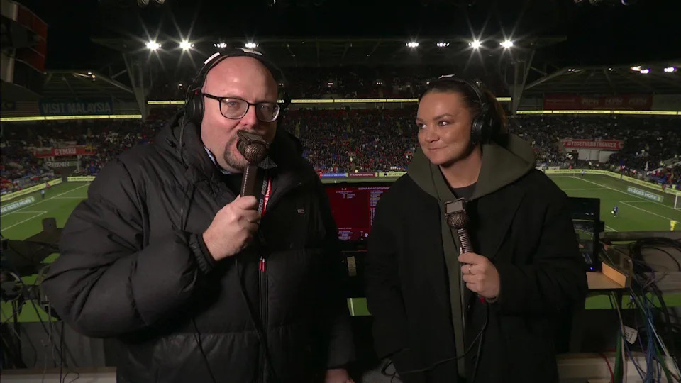 BBC Wales football commentator Simon Davies, holding a microphone in the commentary box, alongside Nia Jones, also holding a microphone - both wearing coats - at half time during the match.