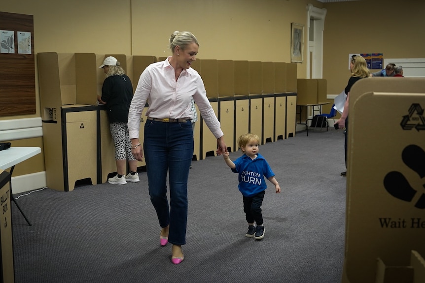 A smiling woman walks while holding the hand of a toddler inside a polling booth