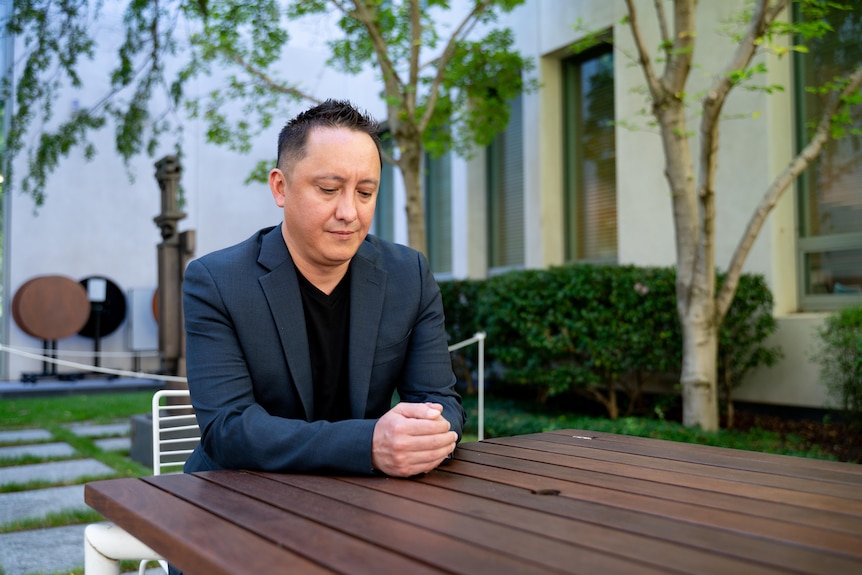 A dark-haired man in a suit sits at an outdoor table and looks at his hands.