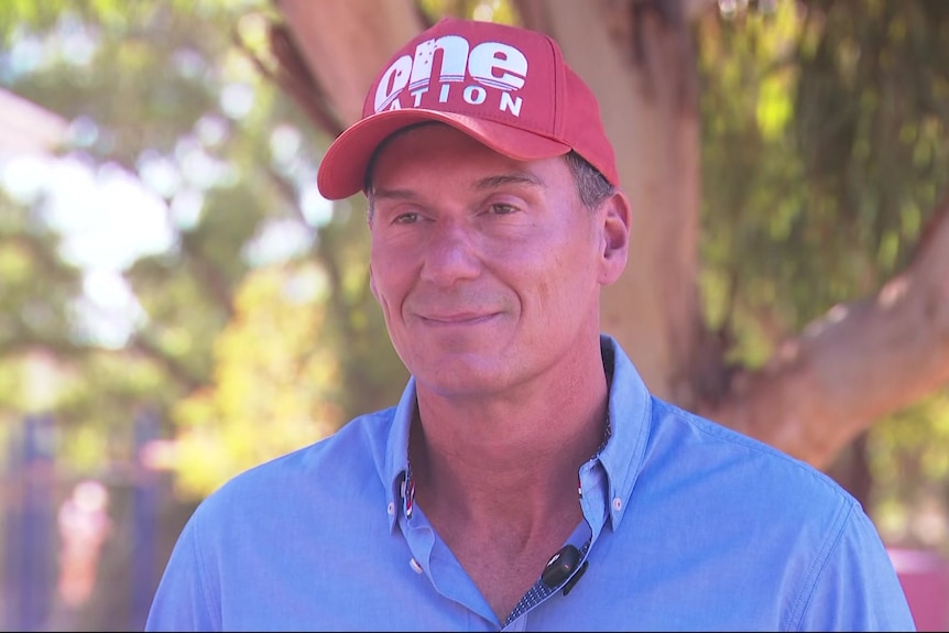 A man standing in a light blue shirt with a red cap with One Nation on it, with trees in the background