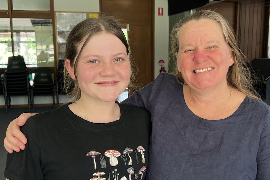 A smiling teenage girl stands beside her mother, who has her arm around her shoulders.