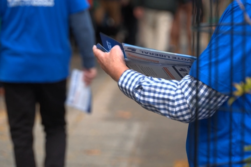 Two unidentifiable people in blue Liberal t-shirts. One is holding a number of how to vote cards with 'Vote Liberal' on them.